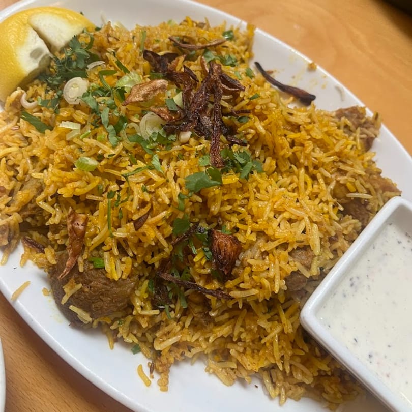 A plate of Indian biryani with rice, meat, fried onions, cilantro, lemon, and a side of raita.