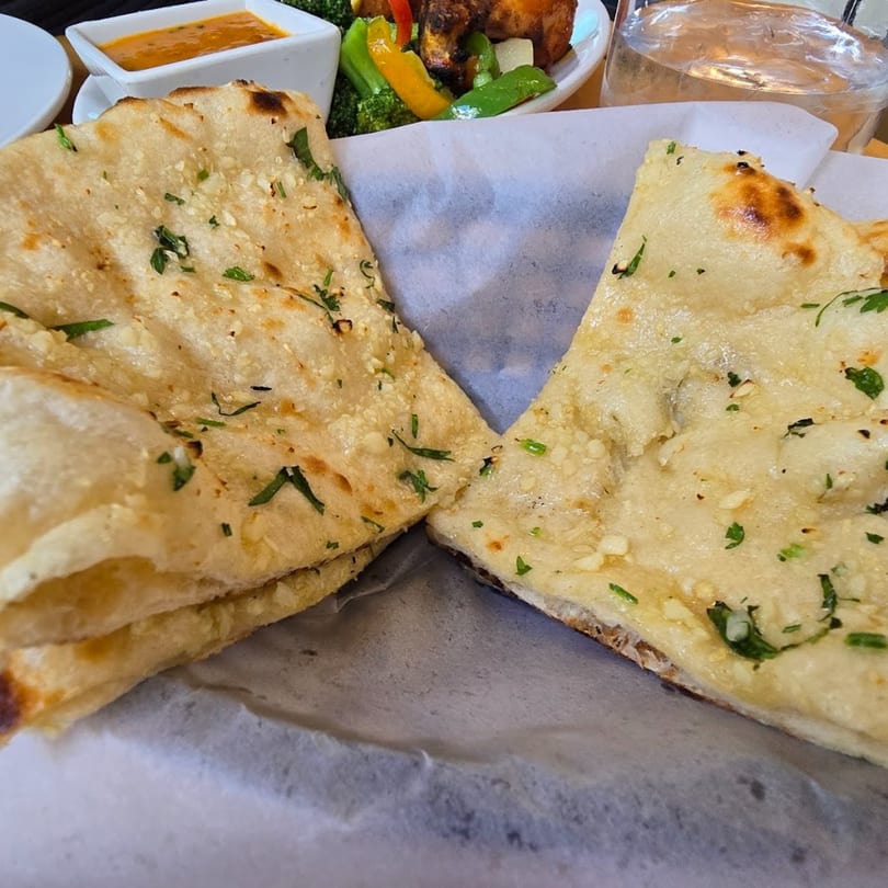 Two pieces of garlic naan bread, garnished with cilantro, served in a basket with Indian dishes in the background.