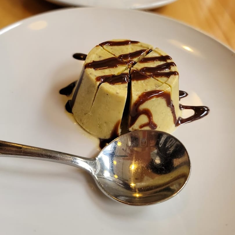 Indian pistachio kulfi dessert, drizzled with chocolate sauce, on a white plate with a spoon.